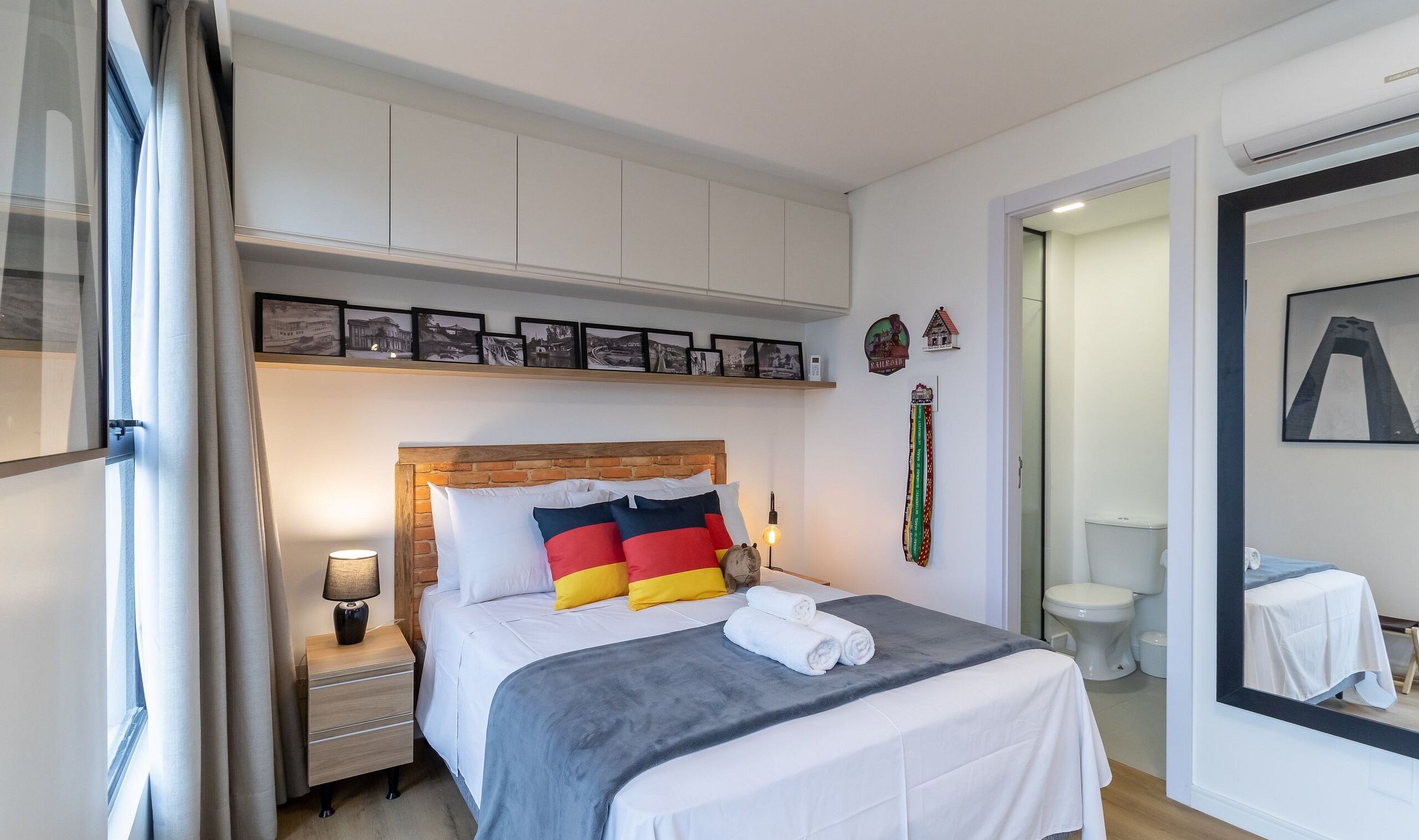 A neatly arranged bedroom with a wooden headboard, a bedside table with a lamp, a bed with colorful pillows, and a shelf with framed pictures.
