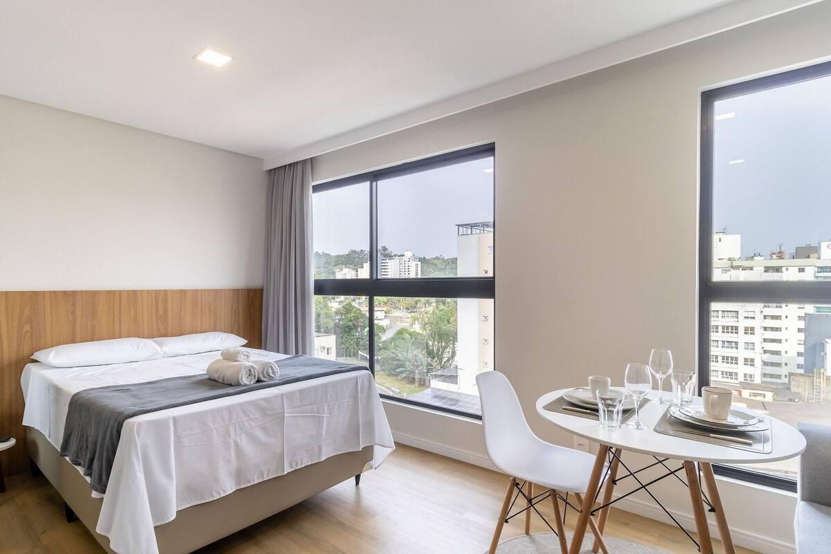A modern hotel room with a bed, a small table with chairs, and large windows offering a city view.