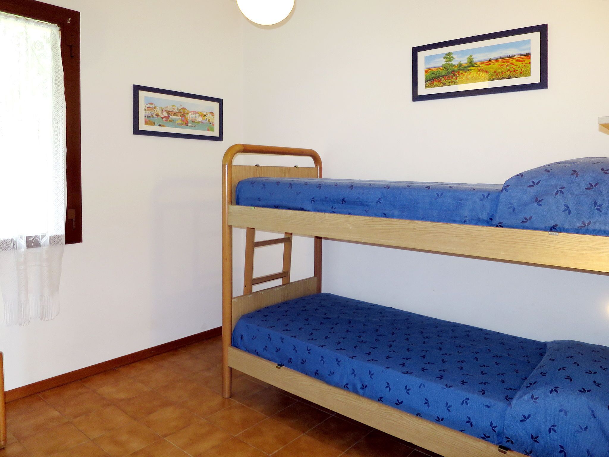 Ein Mehrbettzimmer mit zwei gerahmten Bildern an der Wand, einem Fenster mit Vorhang und einem Fliesenboden.
