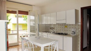 A compact kitchen with white cabinets, a dining table with four chairs, a coffee maker, and a view of a balcony with a yellow wall and white fence. - Villa Stefano by Interhome (Bibione)