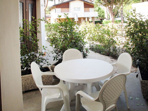 Una mesa redonda con cuatro sillas de plástico blancas en un patio al aire libre, rodeado de follaje y un edificio al fondo.
