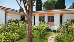 A building with a white exterior and orange trim, featuring a garden with a tree and various plants, and a paved pathway leading to a seating area with white chairs.