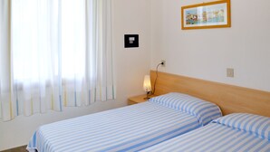 Habitación de hotel con dos camas, una mesita de noche con lámpara, una ventana con cortinas y un cuadro enmarcado en la pared.