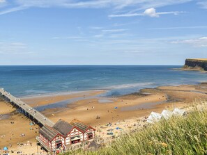 Ein Strand mit einem Steg und Gebäuden mit roten Dächern, mit Blick auf das Meer und die Klippen.