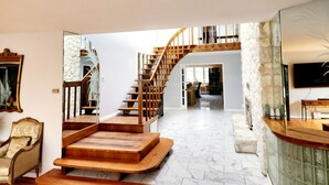 Un'ampia zona giorno con una scala in legno che conduce a un piano superiore, un grande specchio e un televisore su un mobiletto verde.