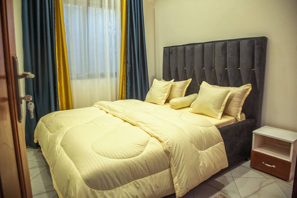 Cama bem arrumada com cabeceira escura, mesa de cabeceira e um pequeno conjunto de gavetas.