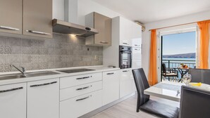Une cuisine moderne avec des armoires blanches, un évier en acier inoxydable et un four encastré. On y trouve un coin repas avec une table et des chaises, et une vue sur la mer grâce à de grandes fenêtres.
