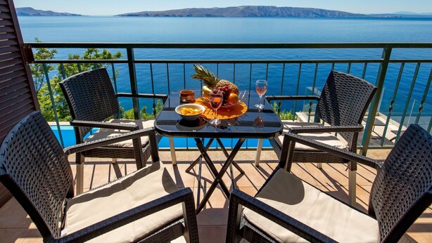 Un balcon agrémenté de fauteuils en osier et d’une table où sont disposés des fruits et des verres à vin, offrant une vue sur une vaste étendue d’eau et des collines lointaines.