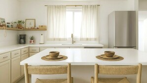 Una cocina moderna con muebles blancos, un refrigerador de acero inoxidable y un comedor con dos sillas tejidas.