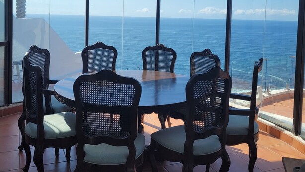 Un comedor con una mesa redonda y seis sillas, ventanas amplias con vista al océano, y un candelabro azul colgante.