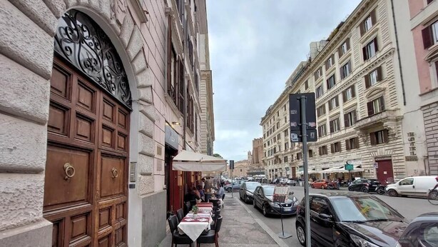Eine schmale Straße mit geparkten Autos und einer Reihe von Tischen und Stühlen im Freien, was auf einen Gastronomiebereich hindeutet.