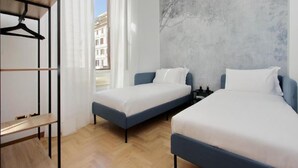 Ein Hotelzimmer mit zwei Einzelbetten, einem Holztisch, einem Kleiderhaken und einem großen Fenster mit Blick auf Gebäude.