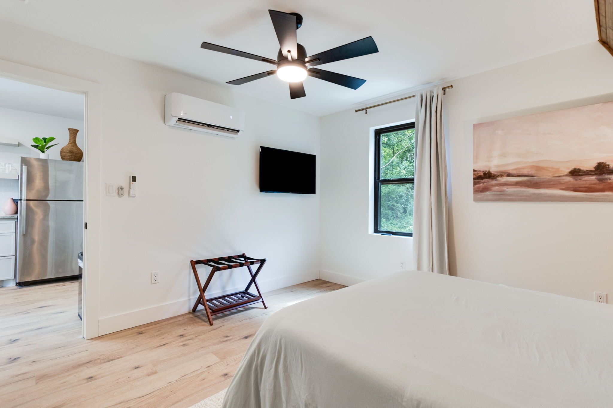 Un dormitorio con cama, ventilador de techo, ventana con cortinas, televisor montado y estante de madera.