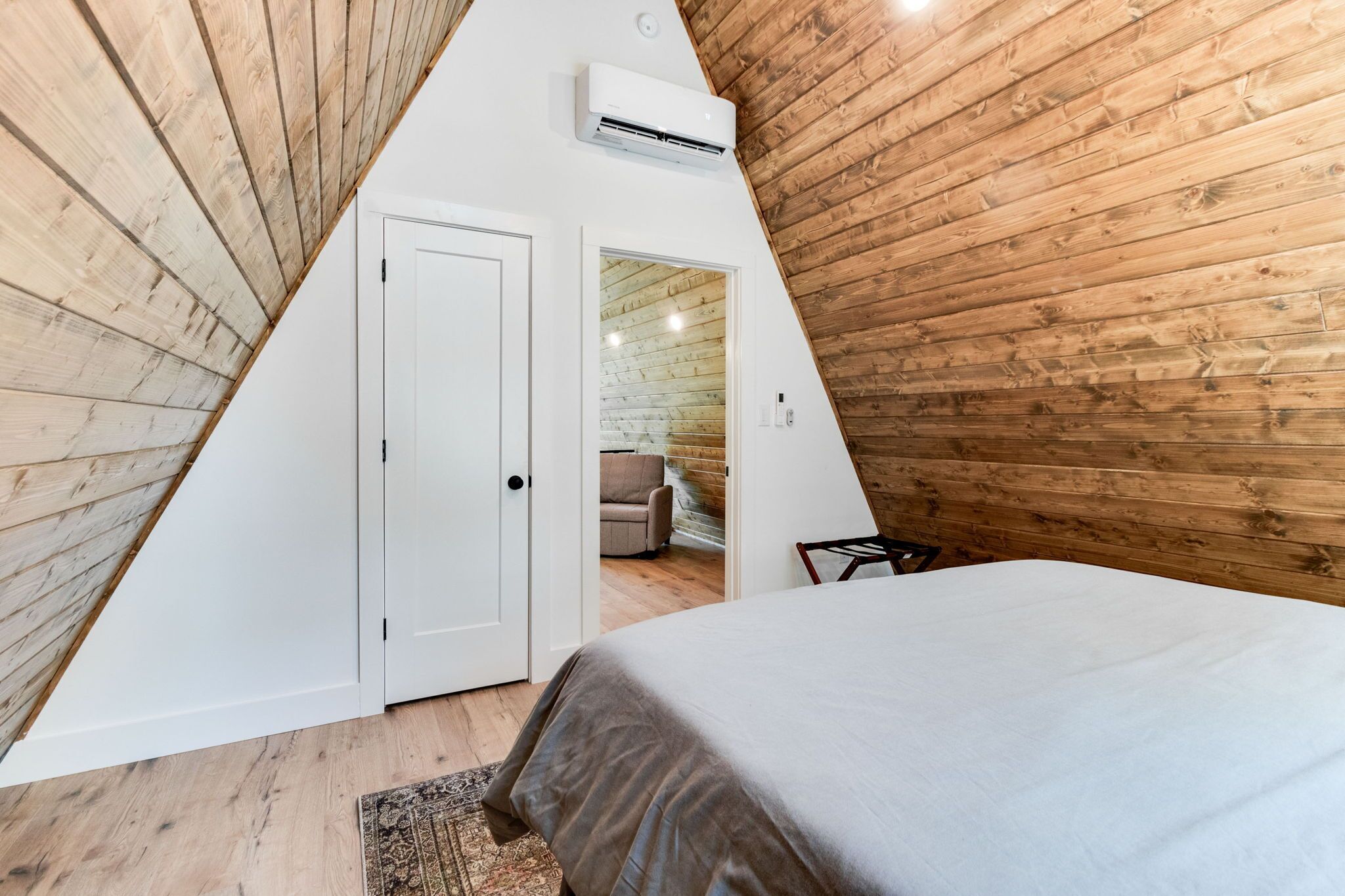 A bedroom with a sloped ceiling, wooden walls, a bed, a door, and an air conditioning unit.