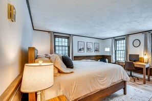 Ein Schlafzimmer mit einem großen Bett, einem Schreibtisch und einem Stuhl. Es gibt Fenster mit Vorhängen, eine Wanduhr und gerahmte Bilder.