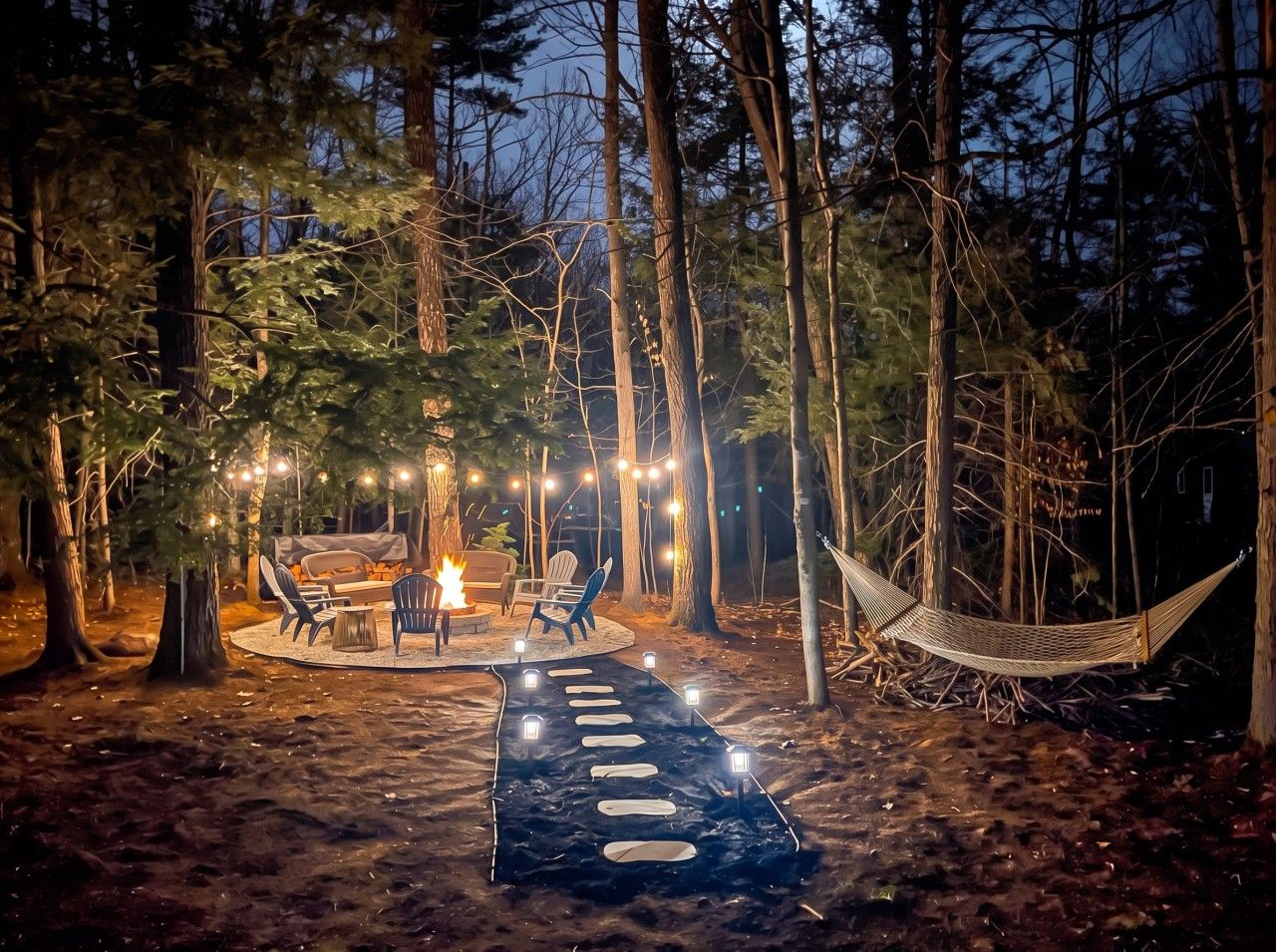En utomhusmiljö under natten, med en eldstad, sittplatser och ljusslingor i en skog.
