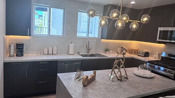 A modern kitchen with dark cabinetry, a central island with bar stools, a stainless steel stove, and pendant lighting. - Spacious Apartment in Prime West Hollywood - Steps from Restaurants & Night life (Los Angeles)
