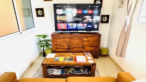 Een woonkamer met een televisie waarop verschillende zenders te zien zijn, een houten kast, een salontafel met boeken en tijdschriften, en een bank.