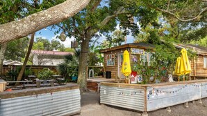 Un espace extérieur aménagé dans un style rustique, avec des tables et des chaises en bois, une barrière en métal ondulé et des parasols jaunes.
