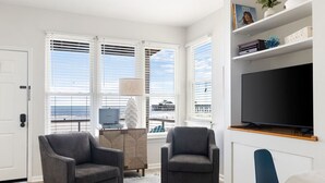 Un salon moderne avec deux fauteuils gris, un téléviseur à écran plat et une vue sur l’océan grâce à de grandes fenêtres.