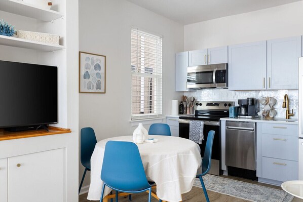 A modern kitchen with white cabinets, a microwave, stove, and dishwasher. There is a dining area with blue chairs and a white table.