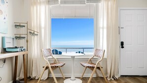 A room with a view of the ocean, featuring a white door, a round table with two chairs, and a desk with a vintage suitcase and a lamp. - Front Beach Condo! 1 Block to Center Street-3C (Folly Beach)