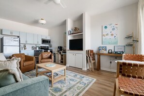 Uma sala de estar moderna com um sofá, mesa de centro e televisão. Há uma cozinha com armários e micro-ondas.