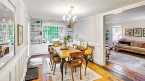 A spacious dining area with a wooden table and chairs, a chandelier, and a view of the outdoors through large windows. - Historic Catskills Farmhouse: Ski, Hike & Relax! (Catskill)