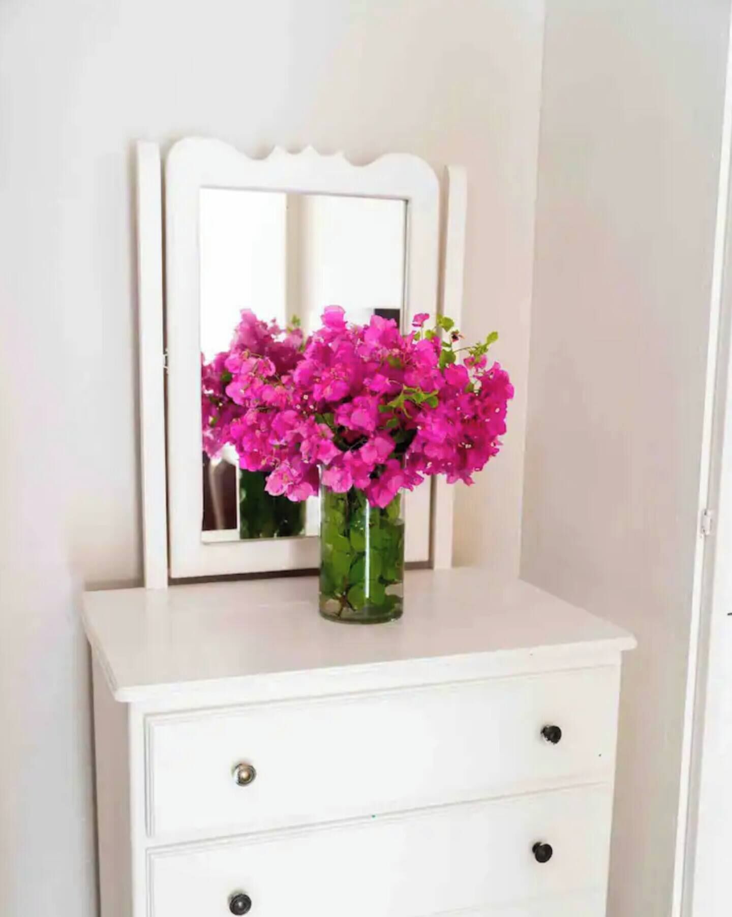 A white dresser with a mirror above it, a vase with pink flowers, and a door to the right.