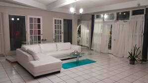 Un salon avec un canapé blanc, une table basse en verre et un tapis bleu. On y trouve une porte avec des panneaux de verre et une plante en pot.
