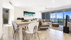 A modern living room with a dining area, a sofa set, and a large window with a view of the ocean.