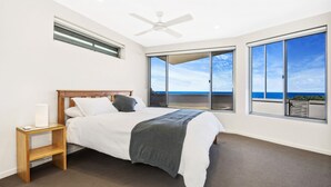 A bedroom with a bed, a nightstand, a ceiling fan, and large windows offering a view of the ocean.