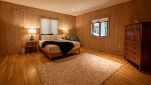 Une chambre avec des lambris en bois, un lit recouvert d’une couette blanche, deux lampes de chevet, une fenêtre avec des stores et une commode en bois.
