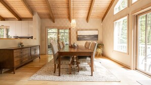 Un vaste espace dédié à la salle à manger, avec une table et des chaises en bois, une grande fenêtre donnant sur la verdure, et une cuisine dotée de meubles et d’un plan de travail en bois.