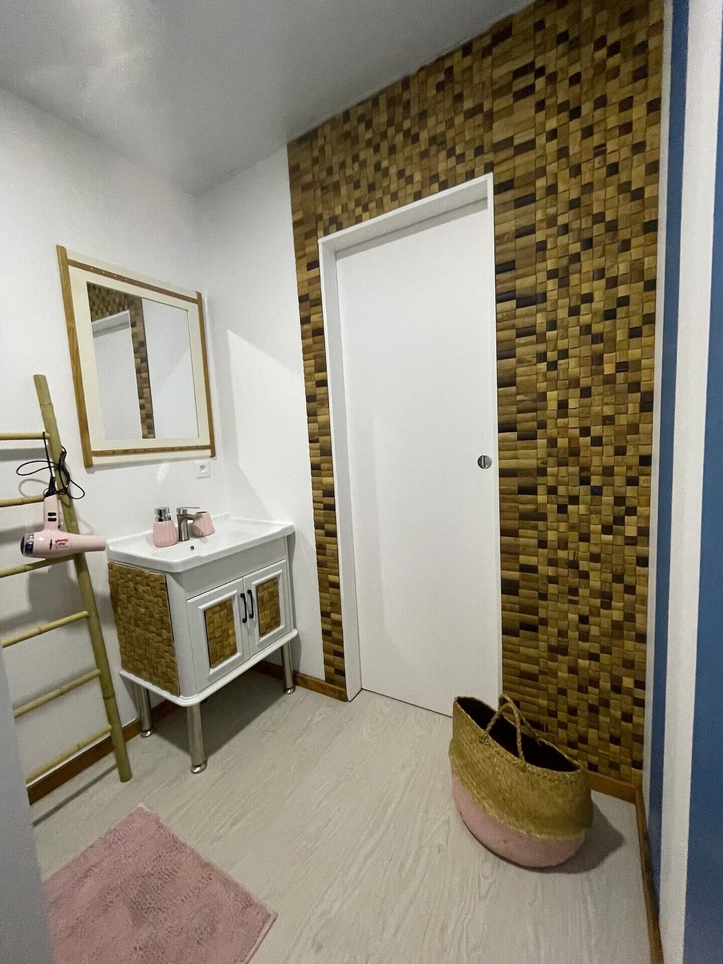 Une salle de bain avec un meuble-lavabo blanc, un miroir, un porte-serviettes et un panneau mural en bois, ainsi qu’une porte.