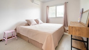 Un dormitorio con cama, un taburete rosa pequeño, un escritorio de madera y una ventana con cortinas.