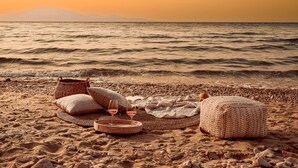 A beach setting with a wicker basket, cushions, a blanket, and a woven ottoman on the sand.