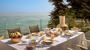 A table set for breakfast with a view of the sea, including fruits, pastries, and beverages.
