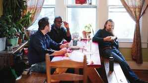 A group of people sitting at a wooden table in a room with large windows, potted plants, and a red curtain. - Bon Paul & Sharky's Youth Hostel (Asheville)