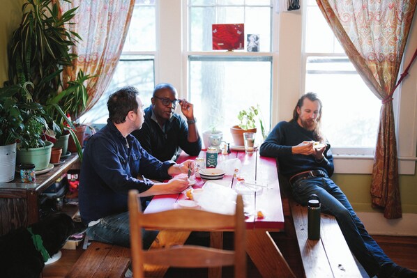 Un grupo de personas sentadas en una mesa de madera en una habitación con ventanas amplias, plantas en macetas y una cortina roja.
