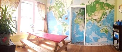 A room with a wooden floor, a red picnic table, a large world map on the wall, a potted plant, and a door leading to another room.