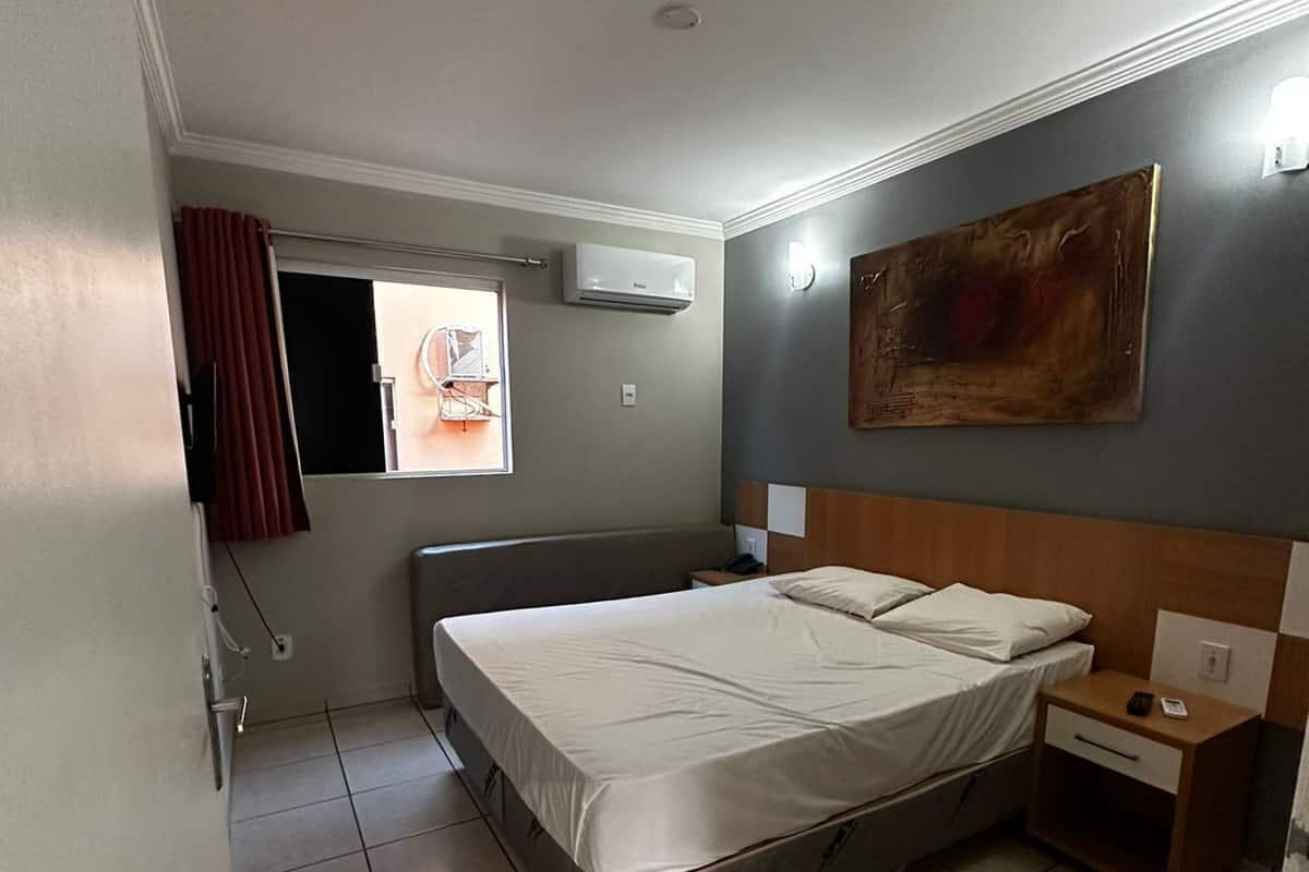 Une chambre d’hôtel avec un lit, une table de chevet, un climatiseur fixé au mur et une fenêtre avec des rideaux.