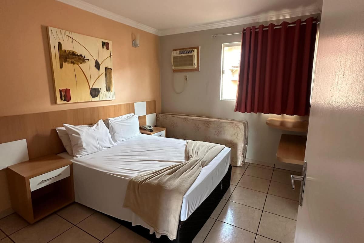 Une chambre d’hôtel avec un grand lit, une table de chevet, un climatiseur fixé au mur, une fenêtre avec des rideaux rouges et une salle de bain avec une douche et un lavabo.