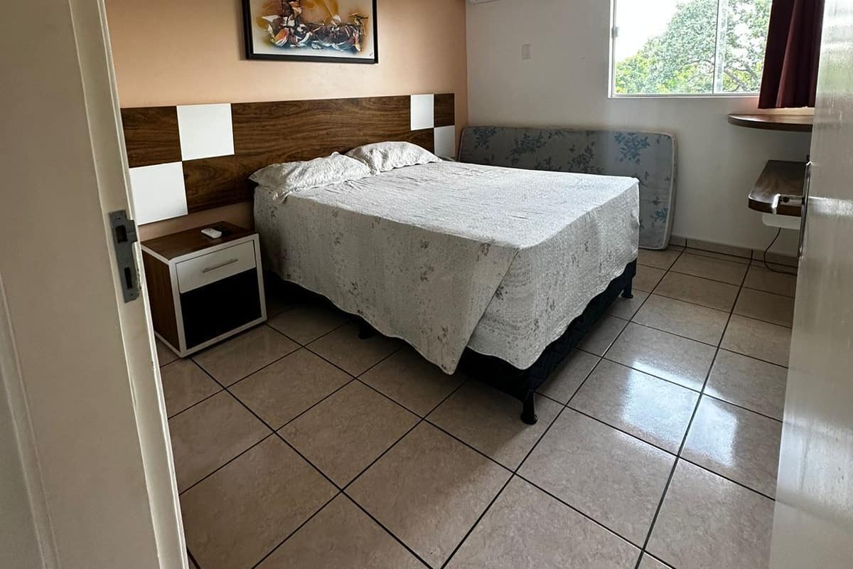 Une chambre à coucher comprenant un lit, une table de chevet, un climatiseur fixé au mur, une fenêtre avec des rideaux et un tableau accroché au mur.