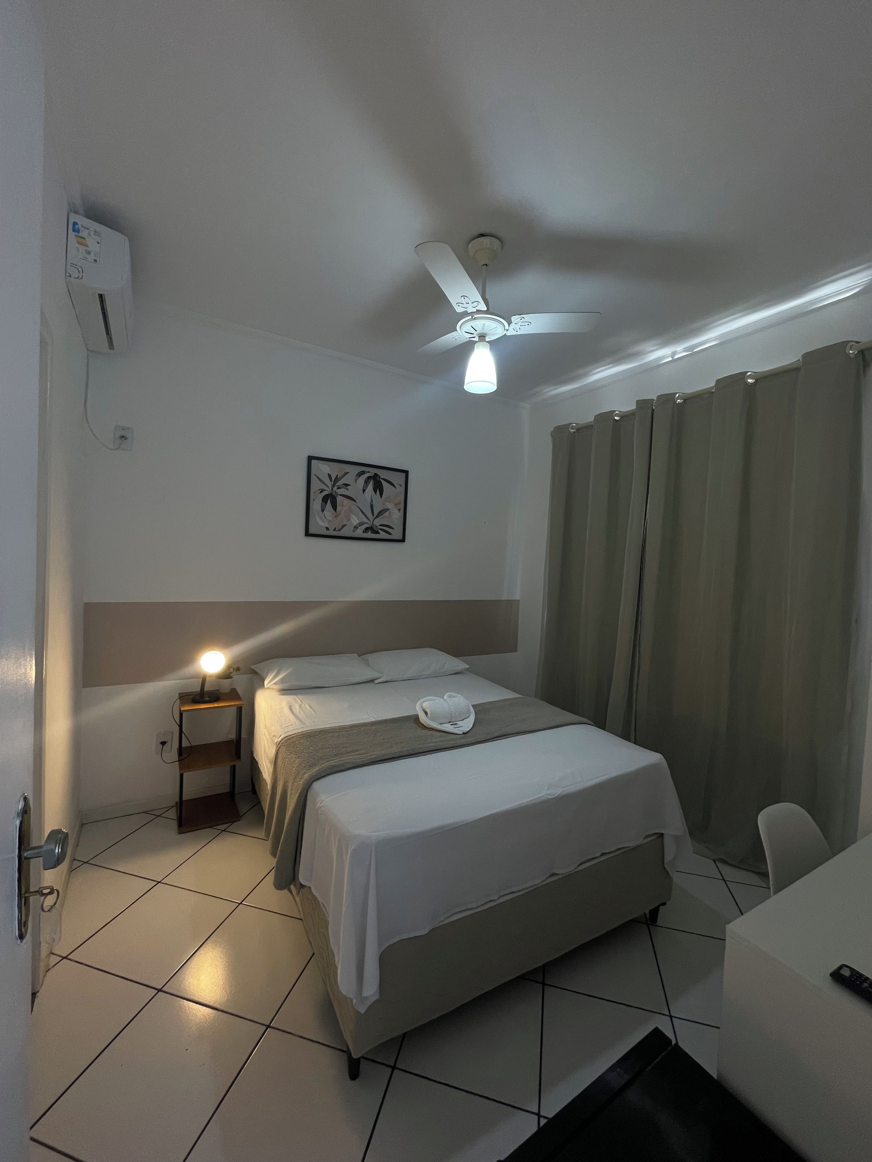 Una habitación de hotel con cama, mesita de noche con lámpara, ventilador de techo, aire acondicionado de pared y ventana con cortinas.