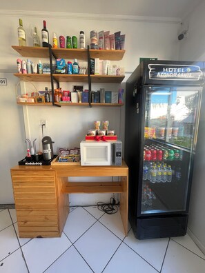 Un minibar de hotel con un refrigerador lleno de bebidas y aperitivos, un microondas y varios artículos de aseo en estantes.