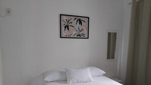 A neatly made bed with white bedding and a green throw blanket, a small bedside table with a plant, and a framed floral artwork on the wall. - Hotel Aconchegante (Jequie)