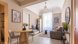 Una sala moderna con una mesa redonda de madera, sillas grises, un sofá gris y un televisor. Hay una estantería y una planta en maceta sobre una consola.