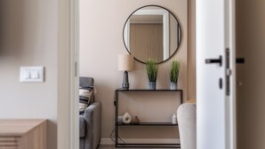 Una sala de estar moderna con un sofá gris, un espejo redondo, una lámpara y una planta pequeña sobre un estante negro.