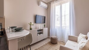 Ein modernes Wohnzimmer mit einem weißen Sofa, einem Essbereich mit weißem Tisch und Stühlen, einem an der Wand befestigten Fernseher und einem großen Fenster mit durchsichtigen Vorhängen.